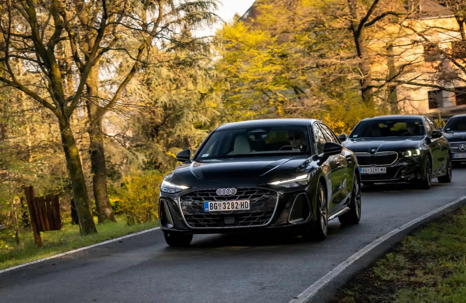 A black Audi leading a BMW and a Mercedes-Benz along a tree-lined road